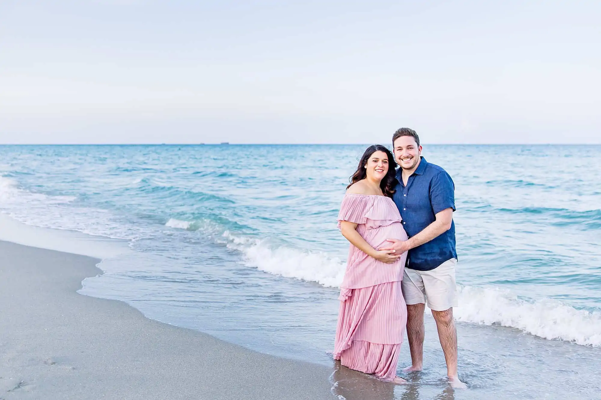 Couple on beach hugging, pregnant mom is wearing a pink tiered ruffled flowy dress - What to Wear for Beach Maternity Photos | Maternity Photographer Miami